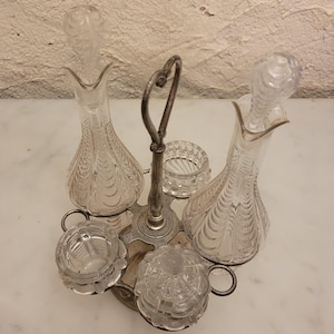 Puede incluir: Un juego de vinagreras de cristal vintage con un soporte de metal plateado. El juego incluye dos decantadores decorativos con tapones, dos platos pequeños abiertos y un plato cubierto. El cristal tiene un diseño texturizado y grabado.