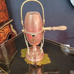 Peut inclure: Une cafetière en cuivre et en laiton avec un corps en cuivre martelé et une poignée en bois. La cafetière est posée sur un support décoratif noir et or.
