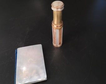 May include: A small, gold-colored, decorative, cylindrical container with a white top and a white and gold striped body. The container is sitting on a black surface next to a small, white, rectangular book with a blue border.
