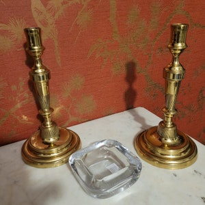 Könnte beinhalten: Zwei goldene Kerzenhalter mit einem durchsichtigen Aschenbecher aus Glas in der Mitte. Die Kerzenhalter haben ein klassisches Design mit einem runden Sockel und einem hohen, schlanken Schaft.