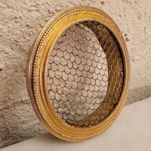 May include: A round, antique brass-colored floral frog with a fine wire mesh center. The rim has a decorative beaded edge and a rope-like design. The floral frog is designed to hold flower stems in place.