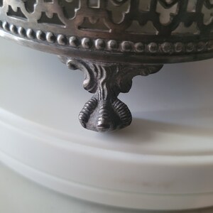 May include: A silver metal base with an ornate design and three claw feet. The base is designed to hold a decorative object.