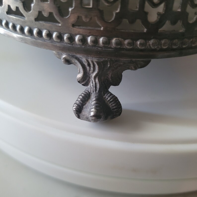 May include: A silver metal base with an ornate design and three claw feet. The base is designed to hold a decorative object.