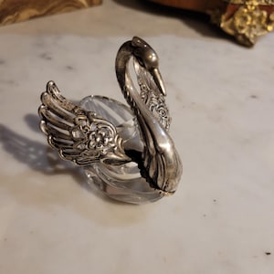 Puede incluir: Un servilletero plateado con forma de cisne, con detalles en las alas y la cabeza. El cisne está posado sobre un soporte de vidrio transparente y redondo. El diseño es vintage y elegante, adecuado para cenas formales.