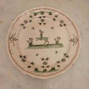 Peut inclure: Une assiette en céramique ronde à bord festonné, dotée d'une base blanc cassé. Le centre présente une illustration peinte à la main en vert représentant un chien debout sur un brin d'herbe, entouré de motifs floraux verts et de fines lignes brunes.