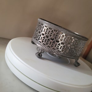 May include: Silver-tone metal decorative stand with an intricate geometric design and three legs. The stand is round and has a raised edge.