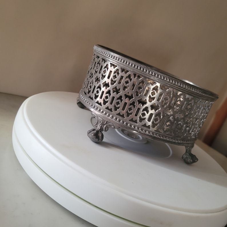 May include: Silver-tone metal decorative stand with an intricate geometric design and three legs. The stand is round and has a raised edge.