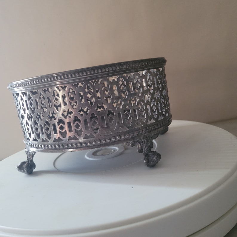 May include: Silver cake stand with an intricate, geometric design and three legs. The stand is made of metal and has a circular shape.
