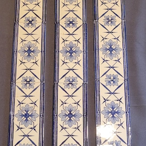 Op de afbeelding: Drie rijen decoratieve keramische tegels met een blauw en wit bloemenpatroon. De tegels zijn verticaal gerangschikt en hebben een crèmekleurige achtergrond.