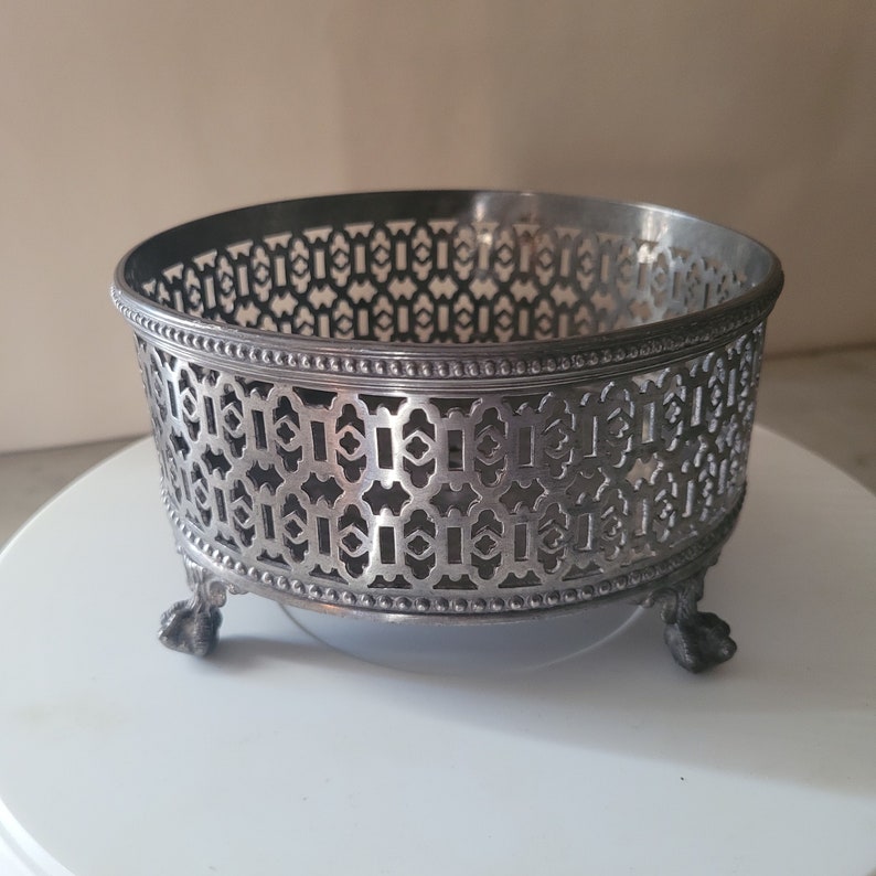 May include: Silver-plated footed bowl with an intricate geometric design. The bowl has a round shape and three legs.
