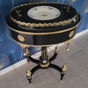 May include: A black and gold oval-shaped table with a decorative top and three legs. The table has a white and blue porcelain plate on top.