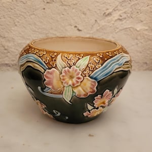 Può includere: Una ciotola in ceramica rotonda con base verde scuro e motivi floreali in rilievo nei toni del rosa, giallo e verde. Il bordo superiore è decorato con un motivo marrone testurizzato e linee ondulate azzurre, con un interno color crema liscio.