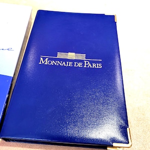 Peut inclure: Un dossier de présentation rectangulaire bleu avec des coins de protection en métal doré. La couverture présente le texte Monnaie de Paris imprimé en doré, surmonté d'un graphique stylisé représentant une façade de bâtiment.