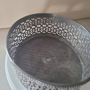 May include: A silver metal cake stand with a decorative, intricate, geometric pattern around the edge. The stand has a circular base and a center hole.