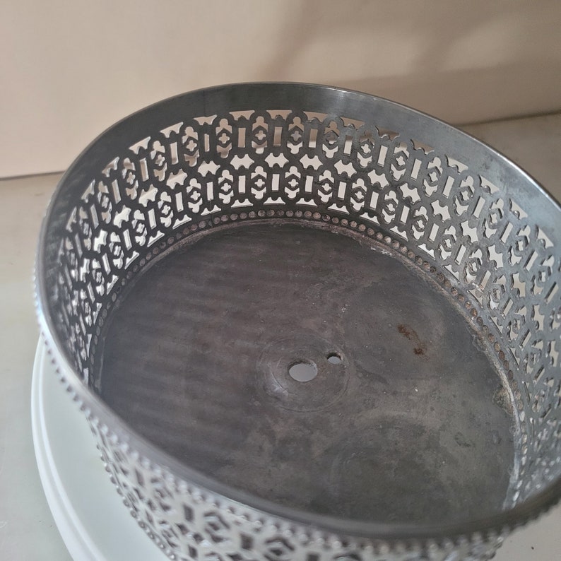 May include: A silver metal cake stand with a decorative, intricate, geometric pattern around the edge. The stand has a circular base and a center hole.