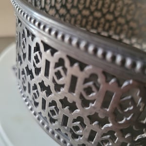 May include: Silver metal cake stand with a decorative, intricate design. The stand has a beaded edge and a circular base.