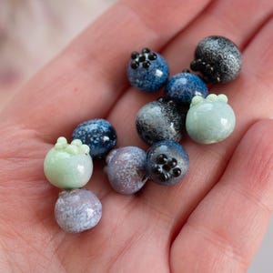 May include: A collection of small, hand-crafted glass beads in shades of blue, green, and white. The beads are shaped like blueberries and have a speckled, textured surface.