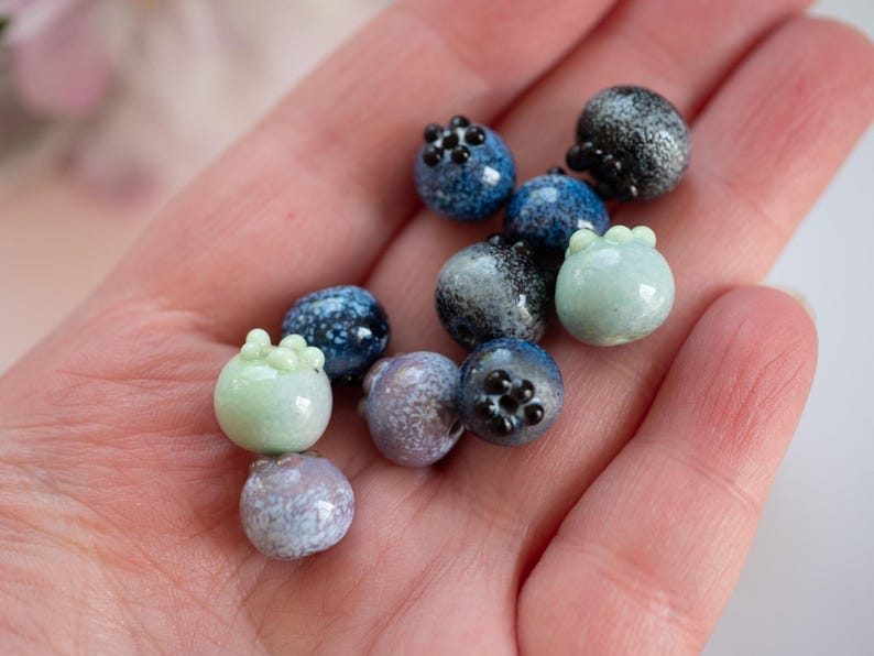 May include: A collection of small, hand-crafted glass beads in shades of blue, green, and white. The beads are shaped like blueberries and have a speckled, textured surface.