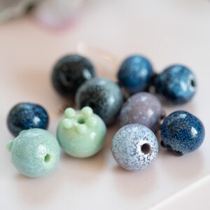 May include: A collection of blue, green, and gray ceramic beads. The beads are round and have a variety of textures and patterns.