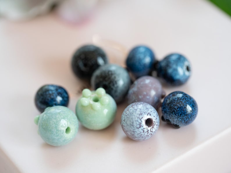 May include: A collection of blue, green, and gray ceramic beads. The beads are round and have a variety of textures and patterns.