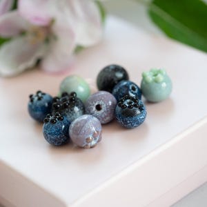 May include: A collection of small, hand-painted glass beads in shades of blue, green, and purple. The beads are shaped like blueberries and have black dots painted on them.