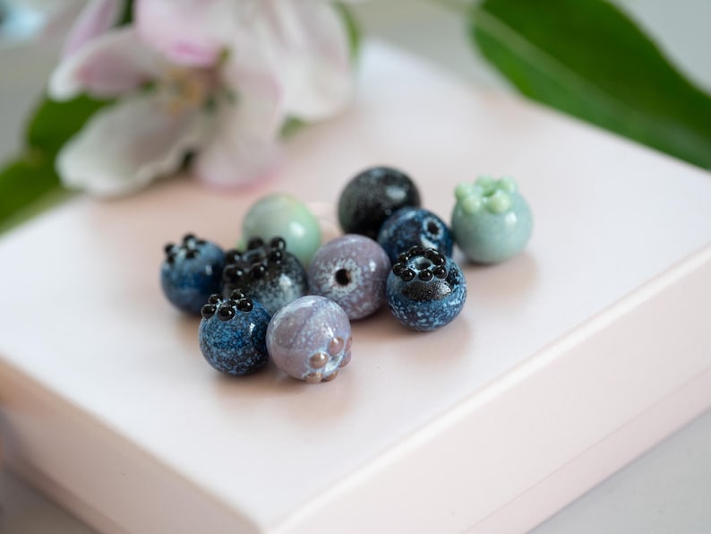 May include: A collection of small, hand-painted glass beads in shades of blue, green, and purple. The beads are shaped like blueberries and have black dots painted on them.