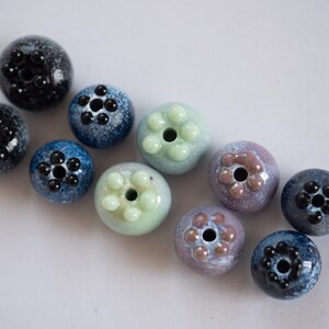 May include: A collection of ten ceramic beads in various shades of blue, green, and purple. Each bead has a unique design with raised dots or a flower-like pattern.