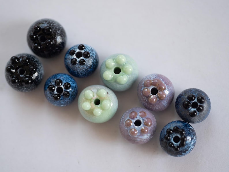 May include: A collection of ten ceramic beads in various shades of blue, green, and purple. Each bead has a unique design with raised dots or a flower-like pattern.