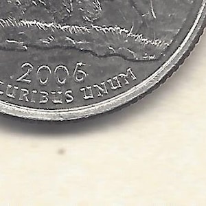 May include: Close-up of a United States quarter coin. The coin is silver and features the year 2006, with the inscription "PLURIBUS UNUM". The coin's edge is ridged.