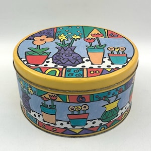 Op de afbeelding: Een ronde gele blik met een kleurrijk ontwerp van potplanten en bloemen. De blik heeft een blauw, groen, rood, oranje en geel ontwerp.