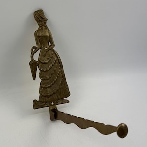 Op de afbeelding: Een vintage messing wandhaak met een gedetailleerde figuur van een vrouw in een lange jurk, die een parasol vasthoudt. De haak strekt zich horizontaal uit met een decoratief golvend ontwerp en een afgerond uiteinde.