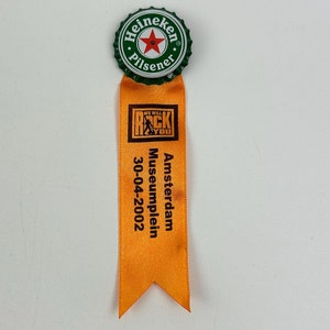 Puede incluir: Un tap&oacute;n de botella Heineken Pilsener verde y blanco con una estrella roja unida a una cinta naranja con el texto "We Will Rock You" y "Amsterdam Museumplein 30-04-2002".