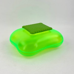 Può includere: Un contenitore di plastica verde brillante con un coperchio verde a forma di biscotto. Il contenitore è progettato per contenere piccoli oggetti.