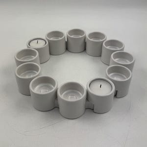 Op de afbeelding: Een cirkelvormige opstelling van twaalf witte keramische kandelaars. Elke houder is cilindrisch met een kleine opening aan de bovenkant. Twee houders bevatten brandende witte kaarsen. De houders zijn verbonden en vormen een doorlopende ring.