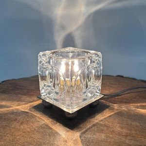 Op de afbeelding: Een heldere glazen kubuslamp met een getextureerd, gegoten ontwerp met afgeronde inkepingen aan elke kant. De lamp wordt van binnenuit verlicht, werpt lichtpatronen en rust op vier kleine, bolvormige metalen voetjes aan de onderkant.