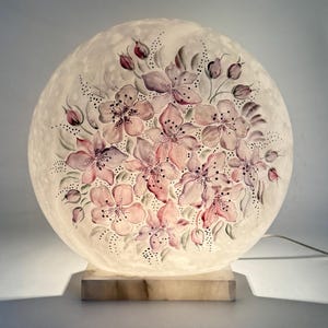 Op de afbeelding: Een ronde lamp met een bloemenontwerp. De lamp heeft een bol met een handgeschilderd patroon van roze en paarse bloemen. Hij staat op een rechthoekige, marmerachtige basis. De lamp is verlicht en geeft een zachte gloed.