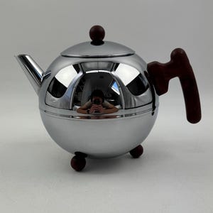 Op de afbeelding: Een verchroomde theepot met een bolvormig lichaam, een kleine tuit en een donkerbruine houten handgreep. Het deksel is bekroond met een kleine, ronde, donkerbruine knop. De theepot staat op drie kleine, donkerbruine voetjes.