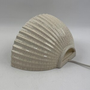 May include: A semi-circular ceramic wall lamp featuring a cream-coloured glaze with a fine crackle finish. The surface is textured with vertical, radiating ridges that fan out from the base. A white electrical cord extends from the side.