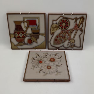 Op de afbeelding: Drie keramische wandtegels met een vintage ontwerp. De eerste tegel heeft een kan, een glas en een perzik. De tweede tegel heeft een vis, een citroen en een vork. De derde tegel heeft drie witte bloemen met oranje centra.