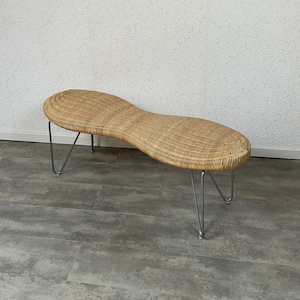 Puede incluir: Un banco de mimbre tejido con una forma curva única y un marco de metal plateado. El banco está sentado en un suelo de baldosas grises.