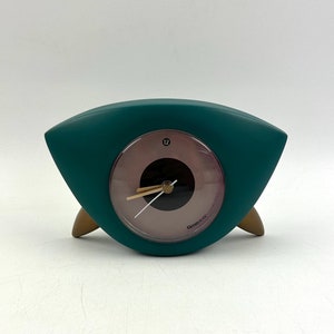 Puede incluir: Reloj analógico verde azulado con detalles dorados y una esfera blanca. El reloj tiene forma de ojo y un pequeño centro negro redondo. La esfera del reloj tiene el número 12 y la palabra "Timex" impresa en ella.