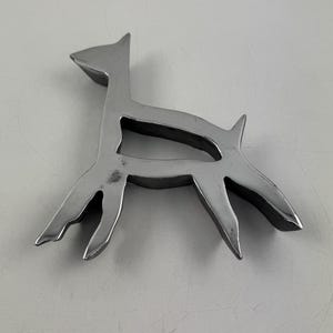 Può includere: Una scultura stilizzata in metallo color argento raffigurante un cane. Il cane è raffigurato in uno stile minimalista, con un contorno semplice e zampe allungate. La scultura è posta su una superficie bianca.