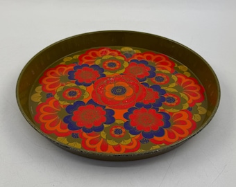 May include: Round metal tray with a floral design. The tray has a dark gold rim and a colorful floral pattern in red, orange, blue, and green. The tray is a vintage piece, perfect for serving or display.