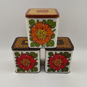Op de afbeelding: Drie vierkante vintage metalen opbergblikken met bloemmotieven. De bovenste blik heeft een bruin deksel met een gouden bloemrand en een grote oranje bloem met een gele kern. De twee onderste blikken hebben witte deksels met een groene bloemrand en een grote rode bloem met een gele kern.