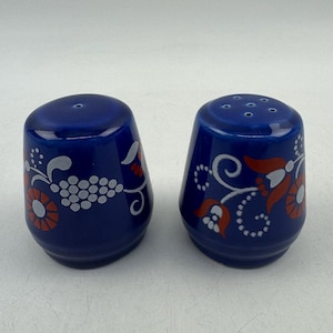 Op de afbeelding: Twee kobaltblauwe keramische zout- en peperstrooiers. Elke strooier heeft een bloemenontwerp in wit en oranje. De zoutstrooier heeft meerdere gaten aan de bovenkant. De strooiers hebben een afgeronde, taps toelopende vorm.