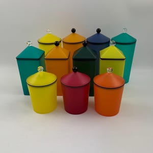 Op de afbeelding: Set van tien kleurrijke glazen potten met deksels. De potten zijn in tinten groen, geel, oranje, rood en blauw. Elke pot heeft een zwarte rand en een doorzichtige glazen knop op het deksel.