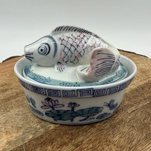 Op de afbeelding: Een keramische sieradenschaal met een visvormig deksel. De vis is wit met roze accenten en zwarte details. De schaal is wit met blauwe bloemen- en geometrische patronen. De schaal staat op een houten oppervlak.