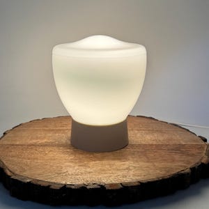 Op de afbeelding: Een tafellamp met een gladde, witte, doorschijnende en afgeronde glazen kap die een zachte gloed uitstraalt. De kap rust op een stevige, cilindrische, lichtgekleurde voet. Een dun wit netsnoer komt uit de voet van de lamp.