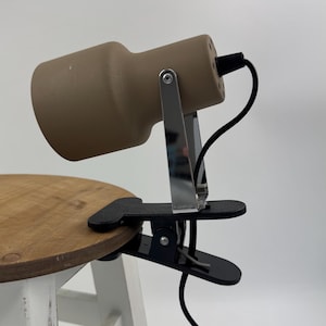 Könnte beinhalten: Ein beigefarbener, zylindrischer Lampenkopf, der an einer silbernen Metallhalterung und einer schwarzen Kunststoff-Federklemme befestigt ist. Die Lampe verfügt über ein schwarzes Stromkabel an der Rückseite des Gehäuses zur Befestigung.