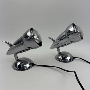 Op de afbeelding: Een paar verchroomde metalen spots met een slank, aerodynamisch ontwerp. Elke lamp heeft een ronde voet, een verstelbare arm en een conische kop met een centrale lamp. Beide eenheden zijn verbonden met zwarte elektrische stroomkabels.